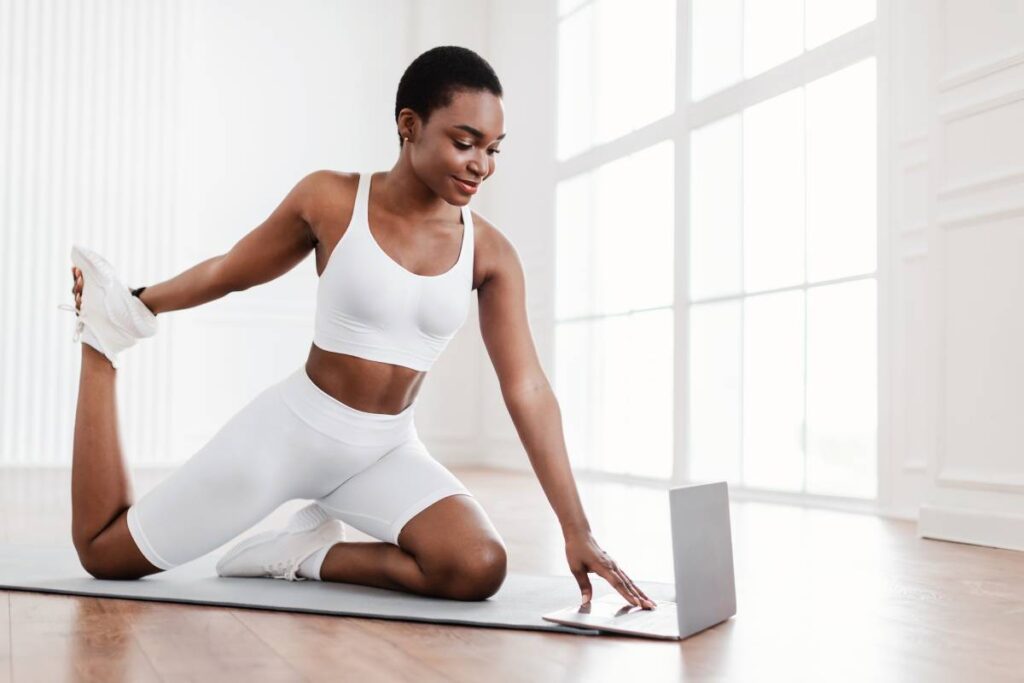 Healthy Sporty Lifestyle. Happy black fit lady in sportswear stretching leg muscles and quads holding feet watching online tutorial on pc exercising in living room or fitness studio, looking at screen