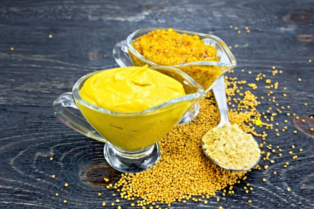 Mustard sauce and Dijon mustard in two glass sauceboats, seeds and powder in a spoon on the background of wooden board