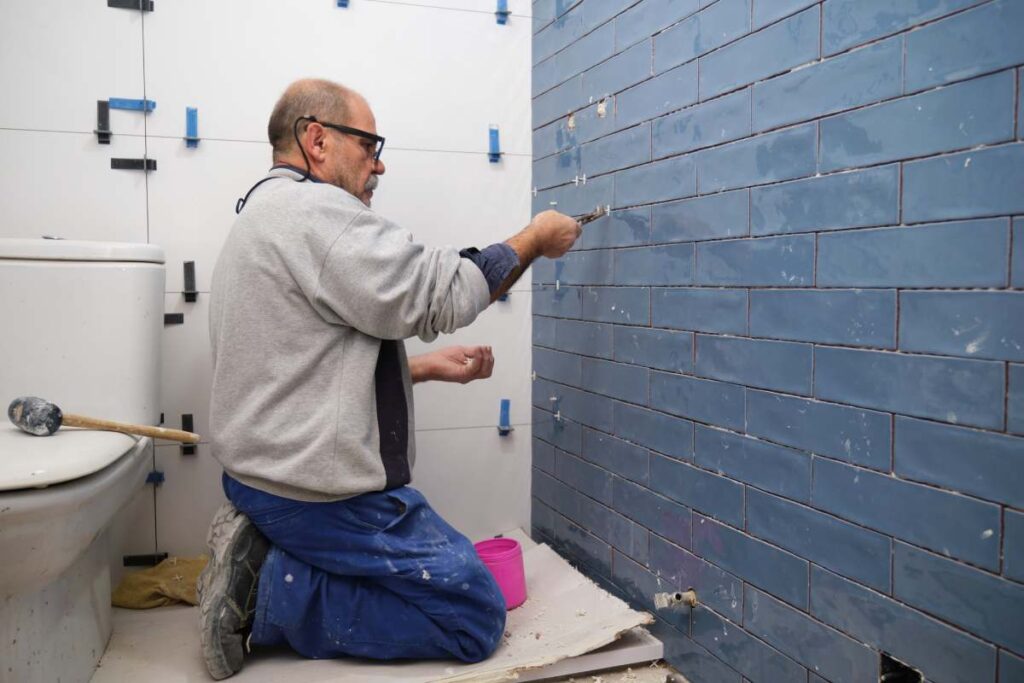 Senior constructor worker removing the separation between blue small ceramic tiles on the wall. Home renovation and building new house concept.