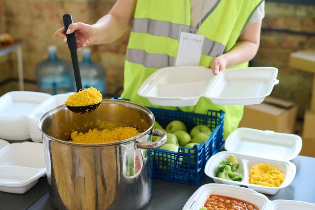 Young female volunteer in uniform holding ladle with cooked food over big pan before putting it into plastic container for refugees