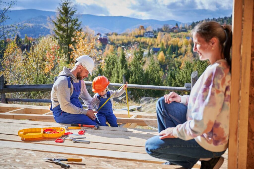 Father, mother and son building wooden frame house. Toddler boy helping his daddy, while woman looking for them on construction site. Guys wearing helmet and blue overalls. Carpentry, family concept.